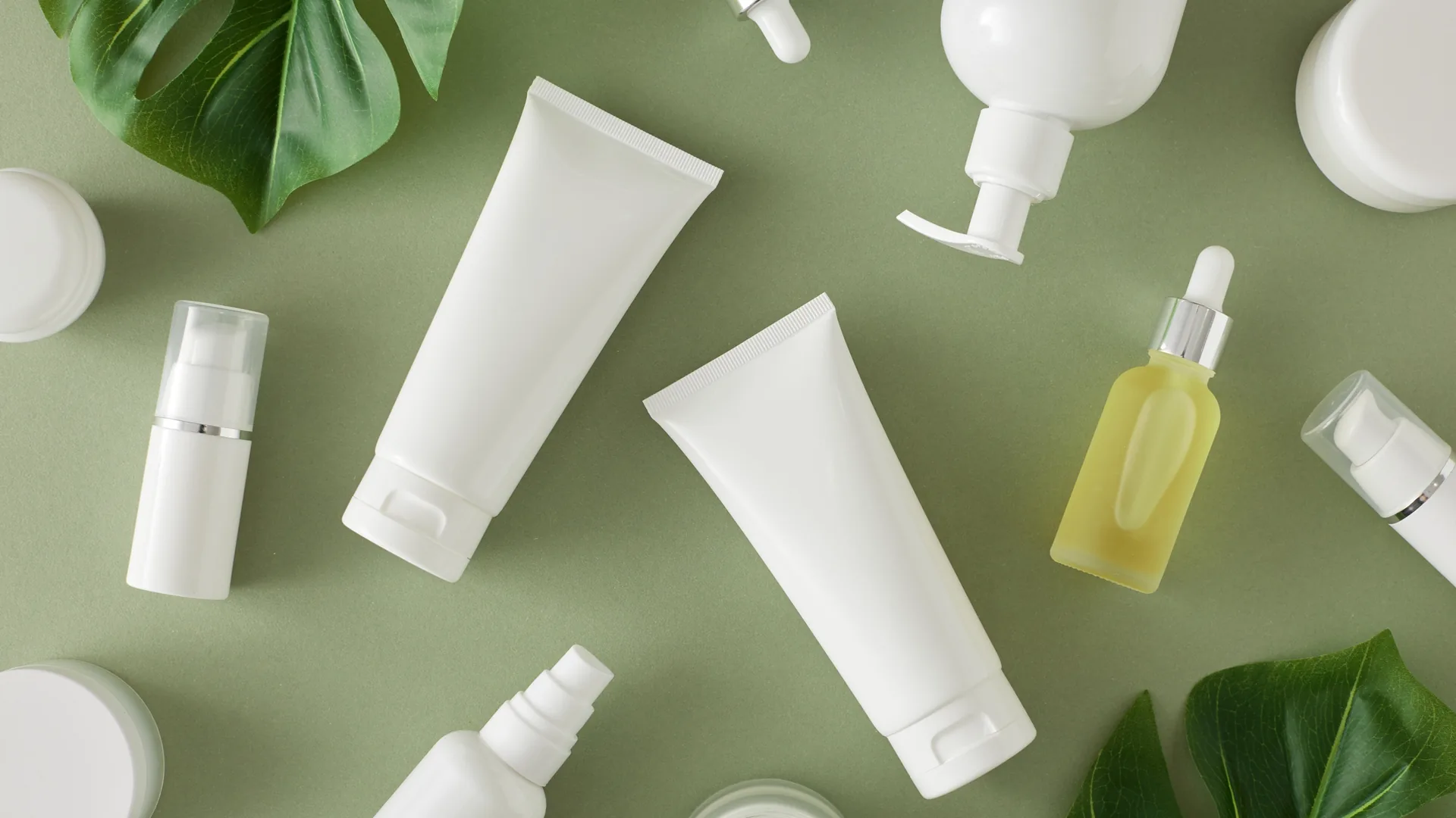 Unbranded white cosmetic tubes, pump bottles, droppers and jars laid flat on a green surface