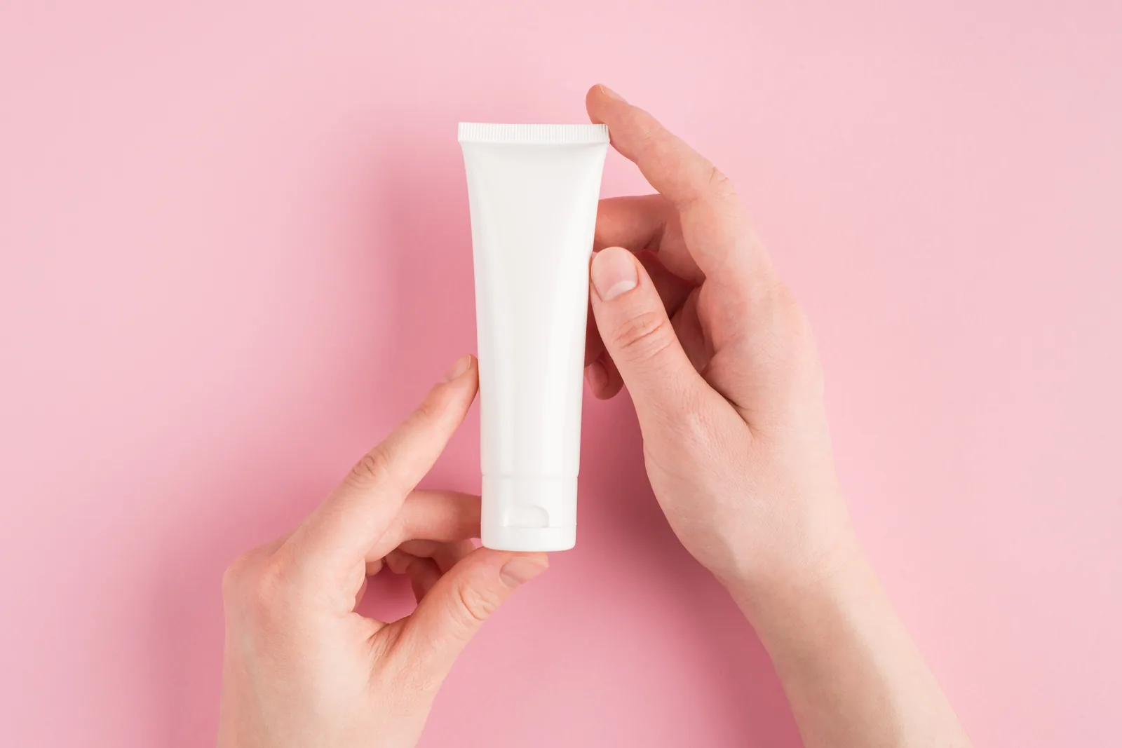 Hands holding an unbranded white cosmetic tube against a pink background