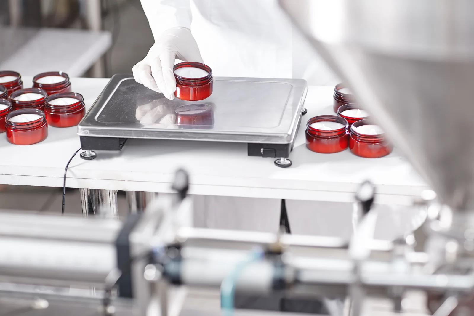 Gloved worker weighing a filled red cosmetic jar on a scale beside a filling machine